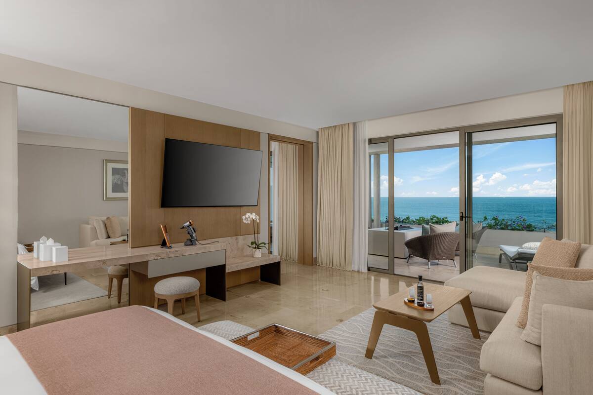 A modern hotel room with a large bed, a desk, a flat-screen TV, a balcony with a view of the ocean, and a seating area with a view.