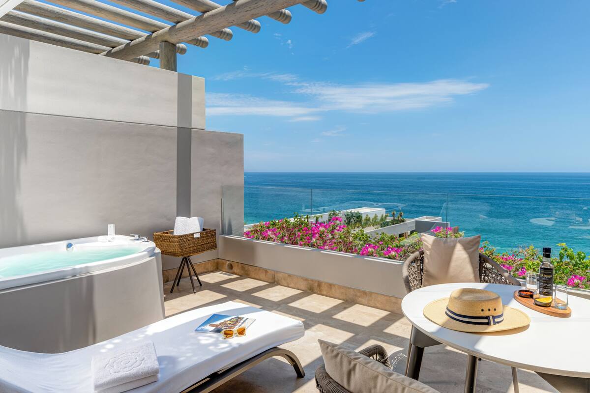 A balcony with a hot tub, a table with a hat and drinks, and a view of the ocean.
