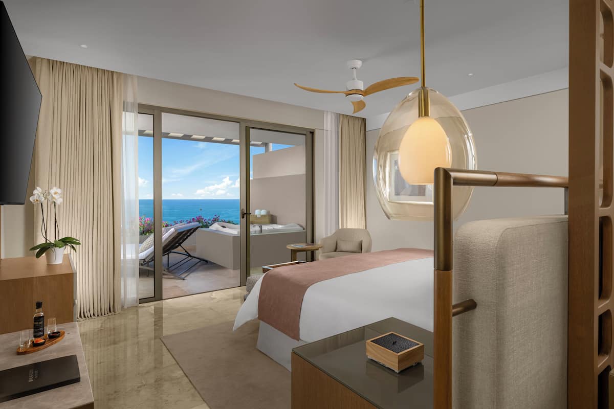 A modern hotel room with a large bed, a desk, a chair, a TV, and a balcony with a view of the sea.