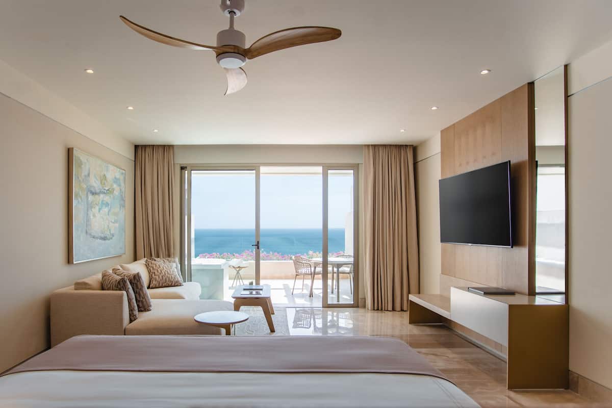 A hotel room with a large bed, a ceiling fan, a TV, a balcony with a view of the ocean, and a dining area with a table and chairs.