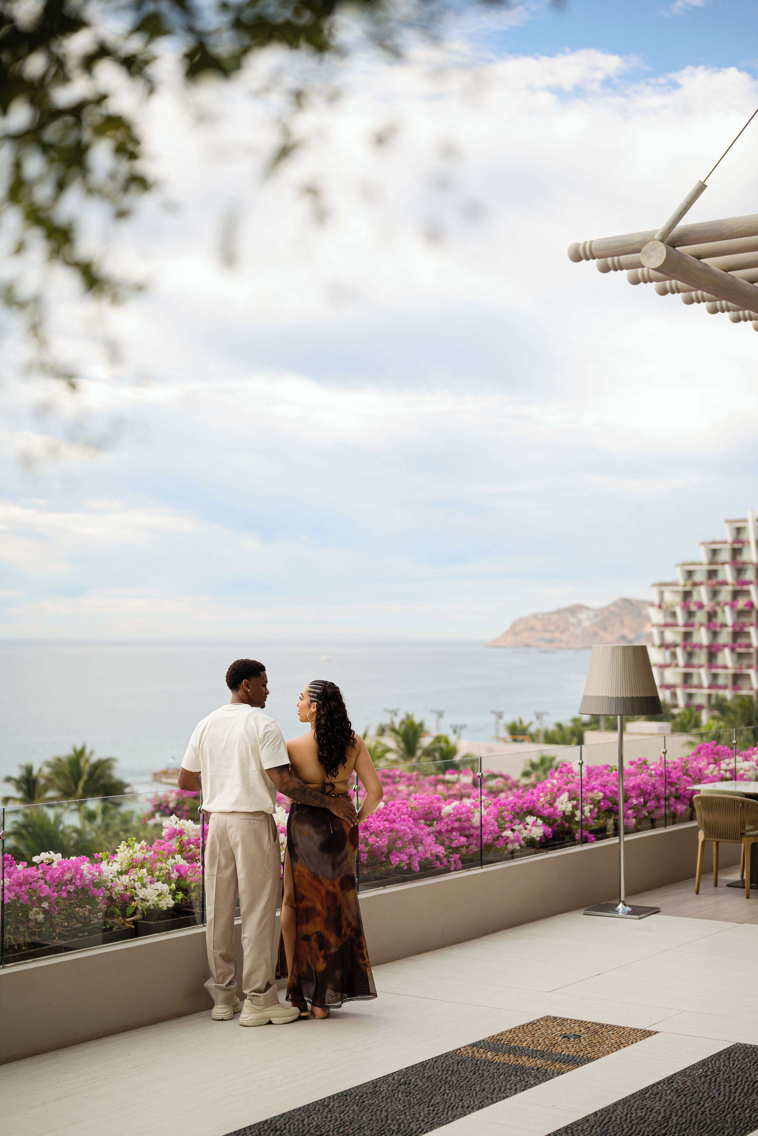 Una pareja está de pie en un balcón con vista a un cuerpo de agua, una montaña distante y un edificio de hotel.
