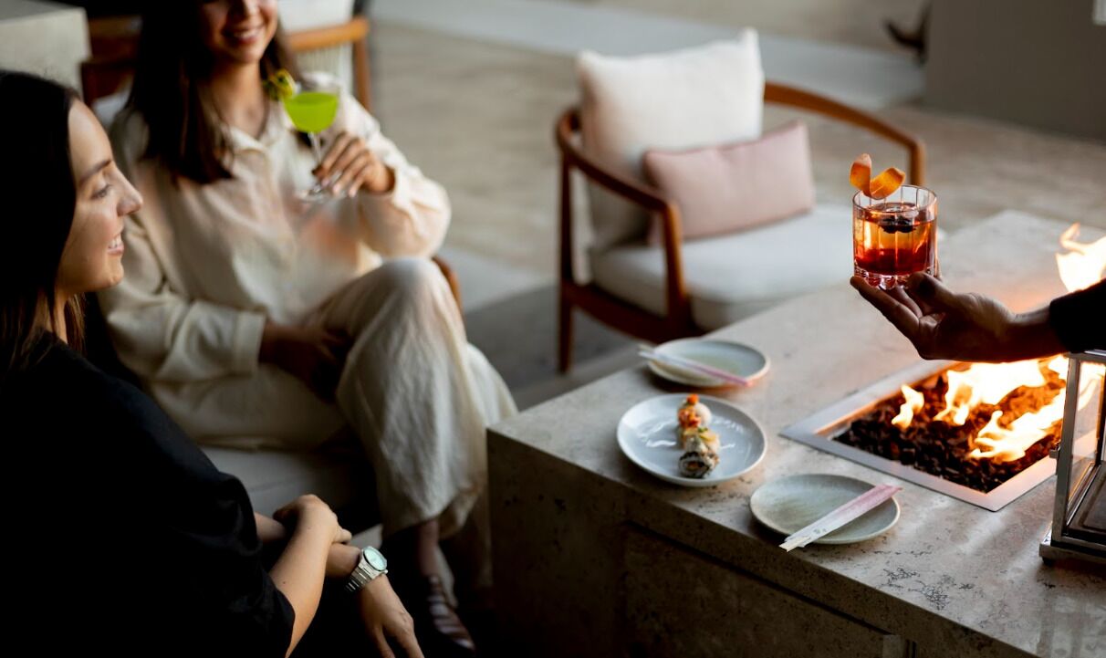 A group of people sitting around a table with drinks and a fire feature.