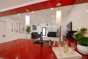 Een moderne keuken met een rood aanrechtblad, roestvrijstalen apparatuur en een eethoek met een witte tafel en wijnglazen.