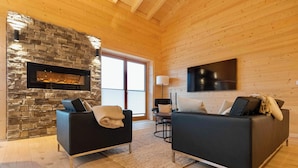 A modern living room with a stone fireplace, two black sofas, a coffee table, and a TV mounted on the wall. - 42 - Bungalow Chalet "Joscha" (Daun)