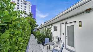 Un corridoio esterno stretto con un vialetto sterrato, un’area relax con tavolo e sedie, e un edificio bianco con una porta e finestre.