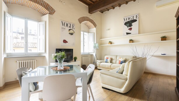 A spacious living room with a glass dining table, white chairs, a beige sofa, and a television. There are two windows with white shutters, a radiator, and a brick archway.