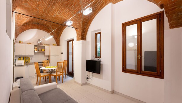 A modern living room with a grey sofa, wooden dining table, and chairs. A TV is mounted on the wall, and there is a kitchen area visible in the background. - San Zanobi 33 - One Bedroom Apartment, Sleeps 2 (Firenze)