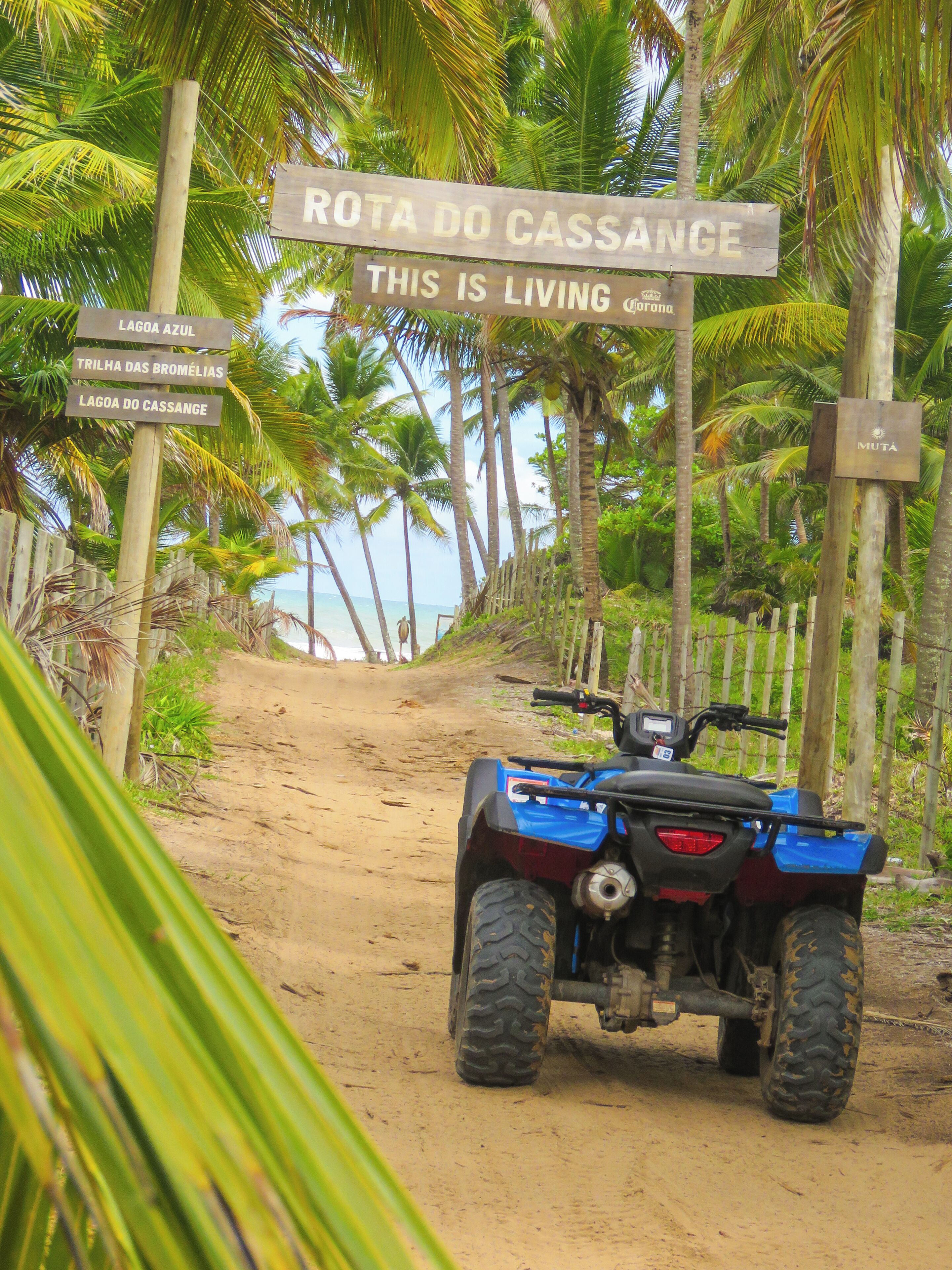 Un sendero de tierra que lleva a una playa, con un cartel que indica 'Rota do Cassange'.