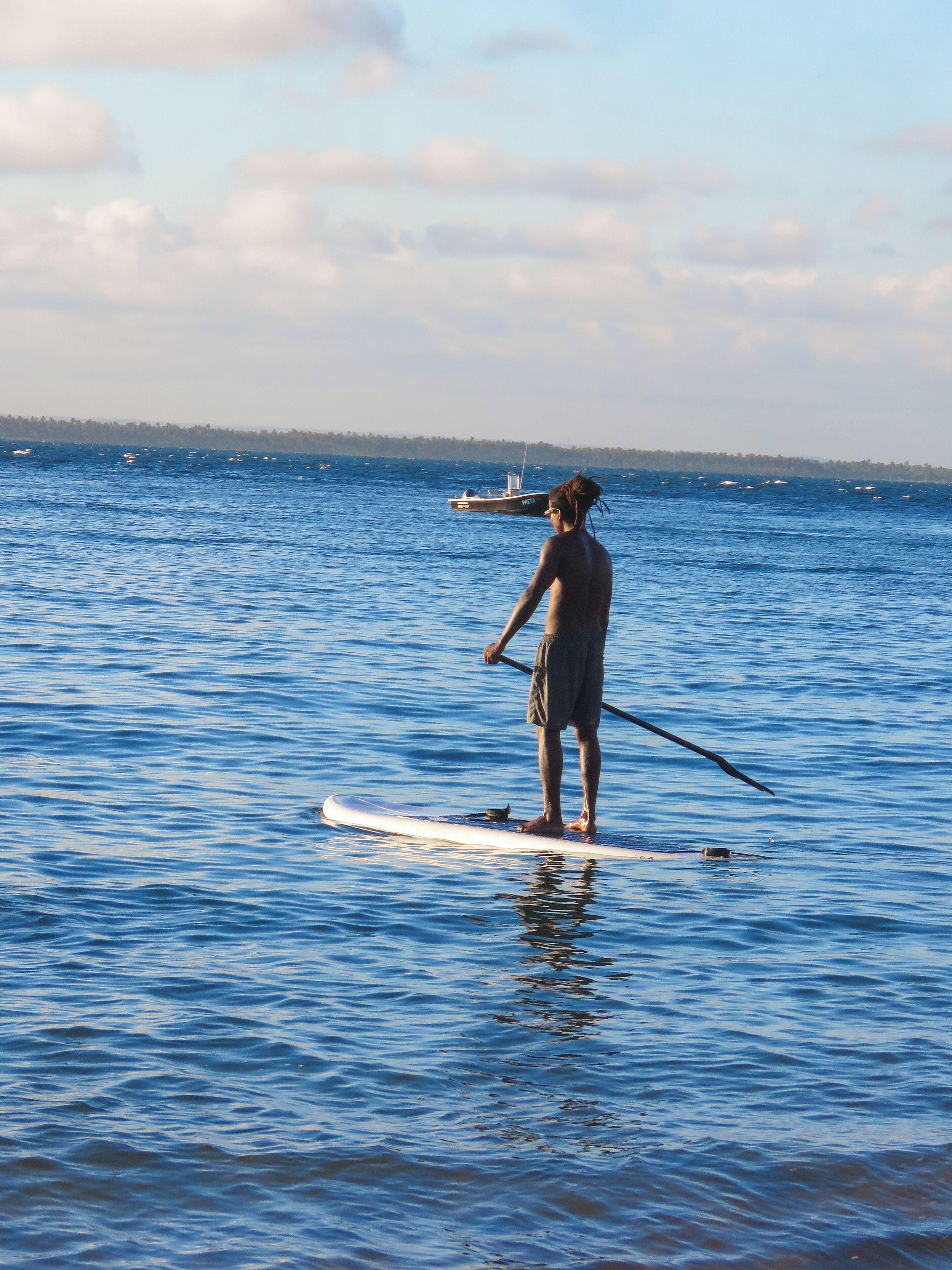 A person paddleboarding on a calm body of water with a distant shoreline and a boat in the background.
