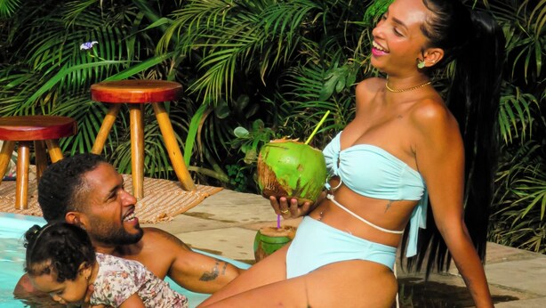 Un área de piscina con una persona sentada al borde, un niño en el agua y un refresco de coco.