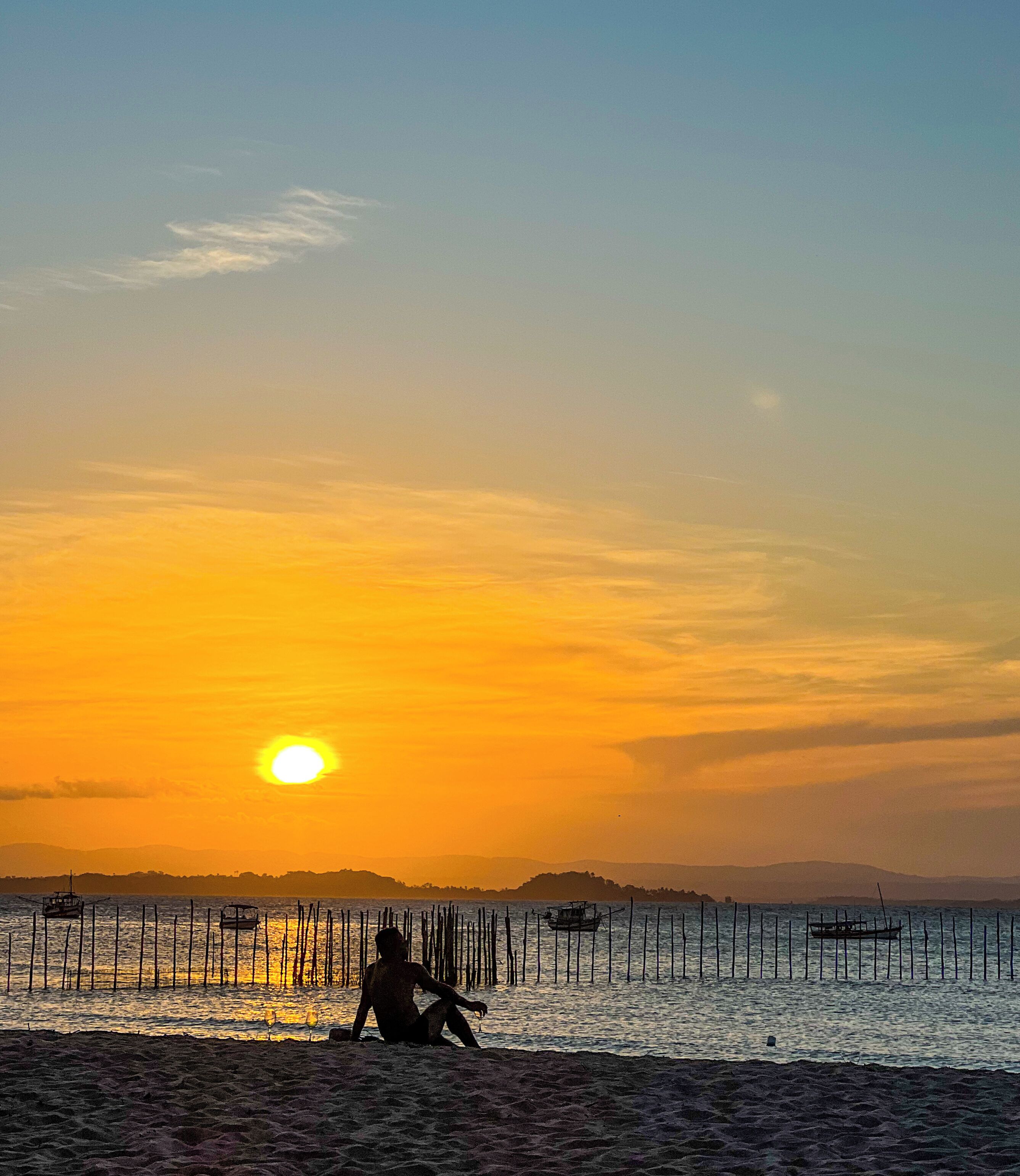 A sunset over a calm sea with a person sitting on the shore.