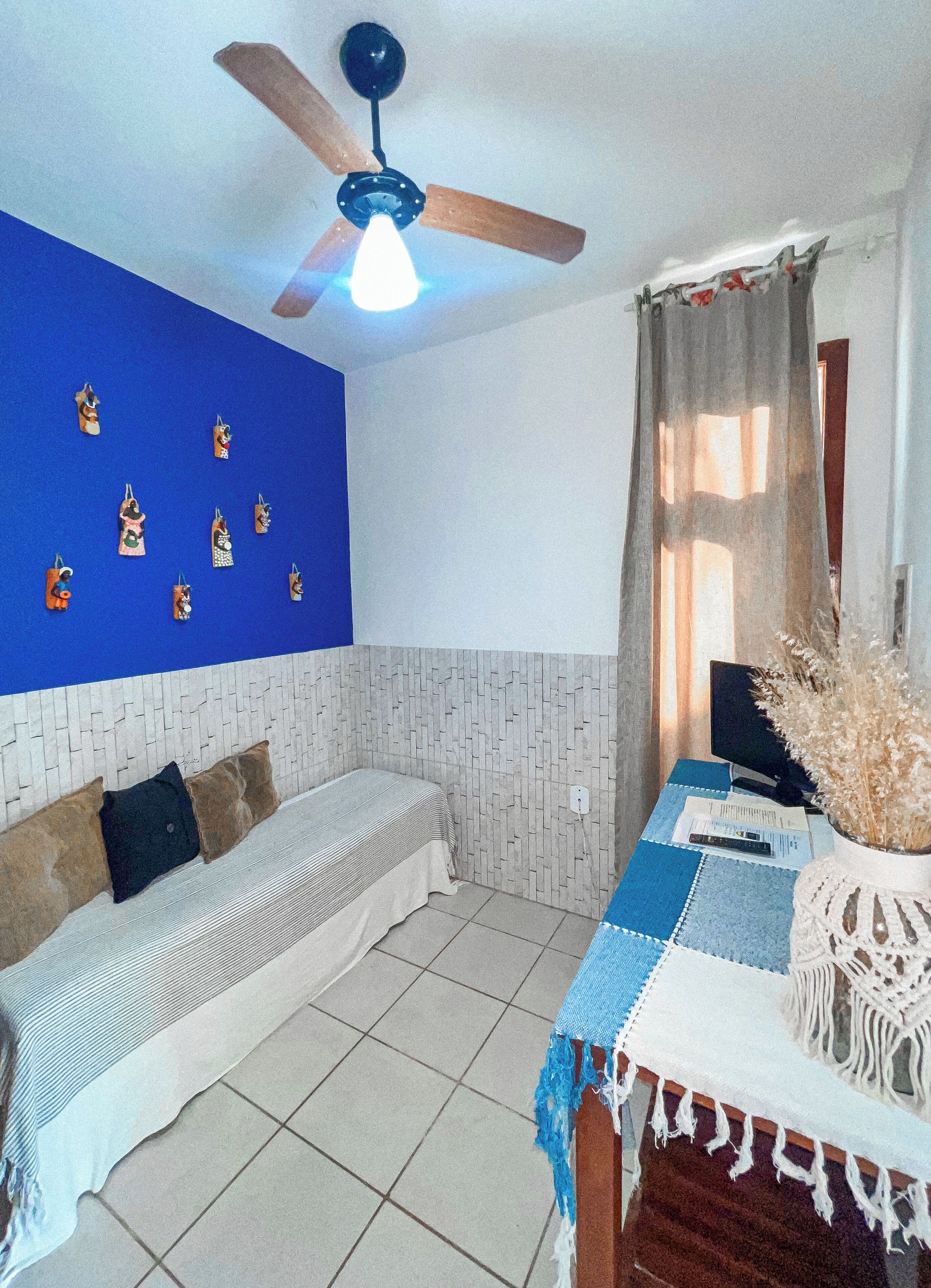 A room with a bed, a table with a laptop, a ceiling fan, and a wall with decorative items.
