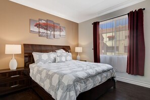 Ein Hotelzimmer mit einem großen Bett, zwei Nachttischlampen, einem hölzernen Kopfteil, einem Fenster mit roten Vorhängen und einem an der Wand hängenden Bild von Bäumen.
