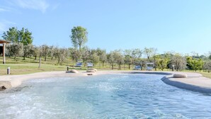 Une piscine aux eaux cristallines, bordée de pierres, entourée d’une pelouse agrémentée d’arbres et comprenant deux espaces de détente à l’ombre.