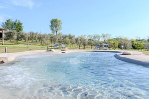 Une piscine aux eaux cristallines, bordée de pierres, entourée d’une pelouse agrémentée d’arbres et comprenant deux espaces de détente à l’ombre.