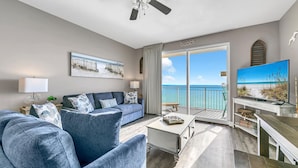 Ein Wohnzimmer mit einer Couch, einem Couchtisch und einem Fernseher auf einem Schrank. Ein Balkon bietet Blick aufs Meer.