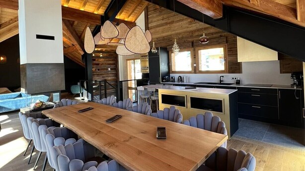 A spacious dining area with a long wooden table and modern chairs, a kitchen with dark cabinetry, and a high wooden ceiling with exposed beams.