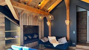 A modern living room with a dark blue sofa, a blue armchair, and a glass coffee table. There are two hanging light fixtures and a wooden ceiling.