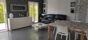 A modern living room with a black leather sofa, a white dining table with chairs, and a kitchen area with a white cabinet and a vase. - Provence house with indoor swimming pool (Pélissanne)