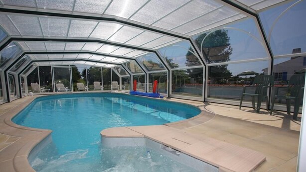 Une piscine couverte avec un jacuzzi, entourée de chaises longues et protégée par une paroi en verre.