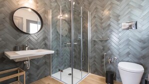 Une salle de bain moderne avec un mur orné d’un motif en chevrons, une douche en verre, un lavabo blanc et des toilettes.