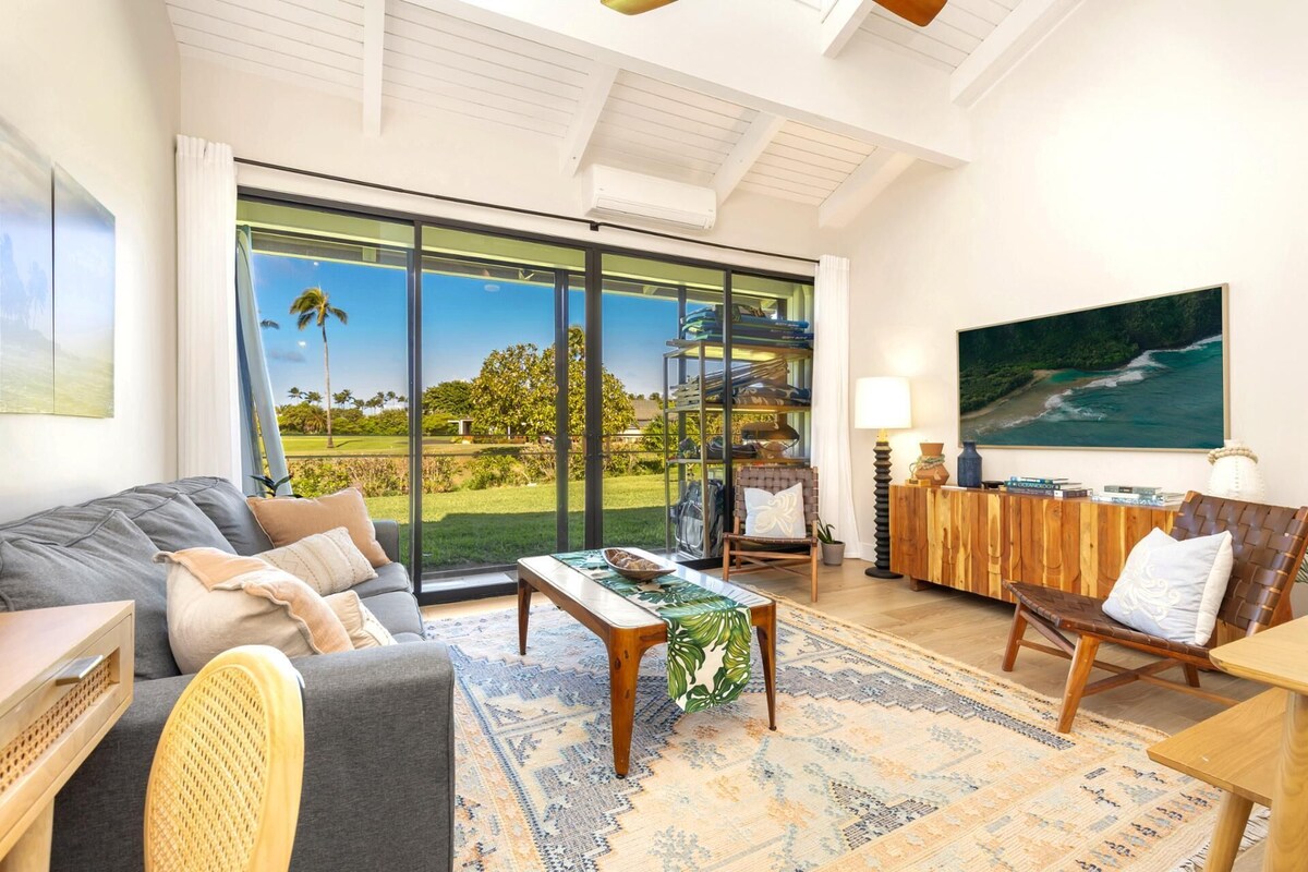 A spacious living room with a large sliding glass door leading to a lush garden, a comfortable sofa, a wooden coffee table, and a bookshelf.