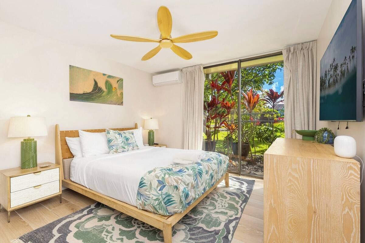 A bedroom with a bed, bedside tables, a ceiling fan, a painting, a window with curtains, and a view of greenery outside.