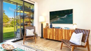 Een kamer met een glazen deur naar een zwembadgedeelte, twee comfortabele fauteuils, een houten bijzettafel met decoratieve objecten, en een groot schilderij met uitzicht op de oceaan.