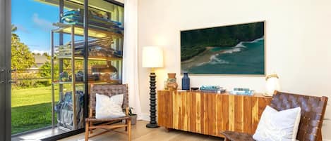 Una habitación con puerta de cristal que da a una zona de piscina, dos sillones cómodos, una mesa consola de madera con objetos decorativos y un cuadro de una amplia vista al océano.