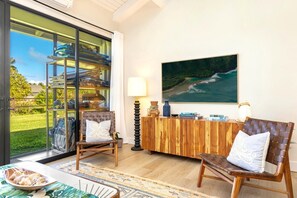Ein Zimmer mit Glastür, die zu einem Poolbereich führt, zwei gepolsterte Sessel, ein Holzkonsolentisch mit Dekoration und ein großes Gemälde mit Meerblick.