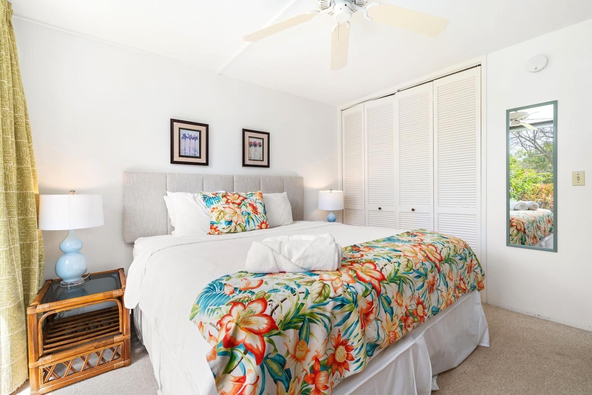 A bedroom with a bed, two bedside lamps, a ceiling fan, a window with a view, and a mirror.