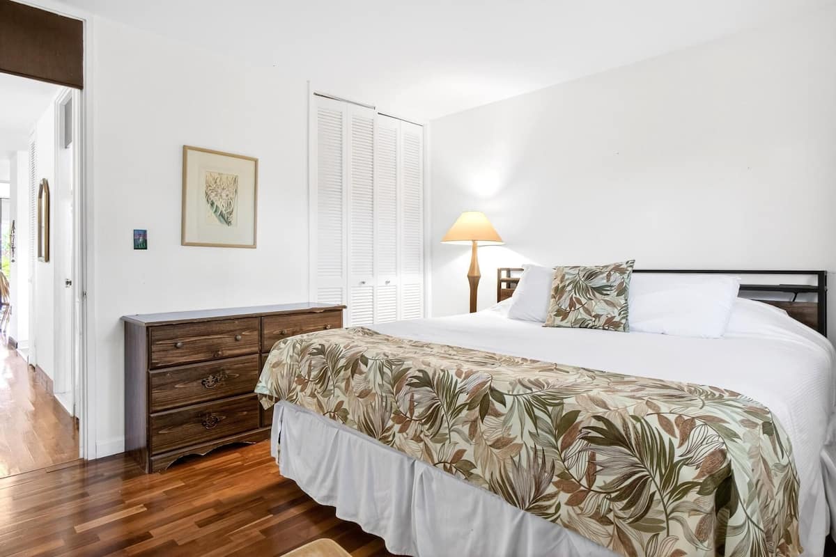 A bedroom with a bed, a wooden dresser, a nightstand with a lamp, and a door with white shutters.