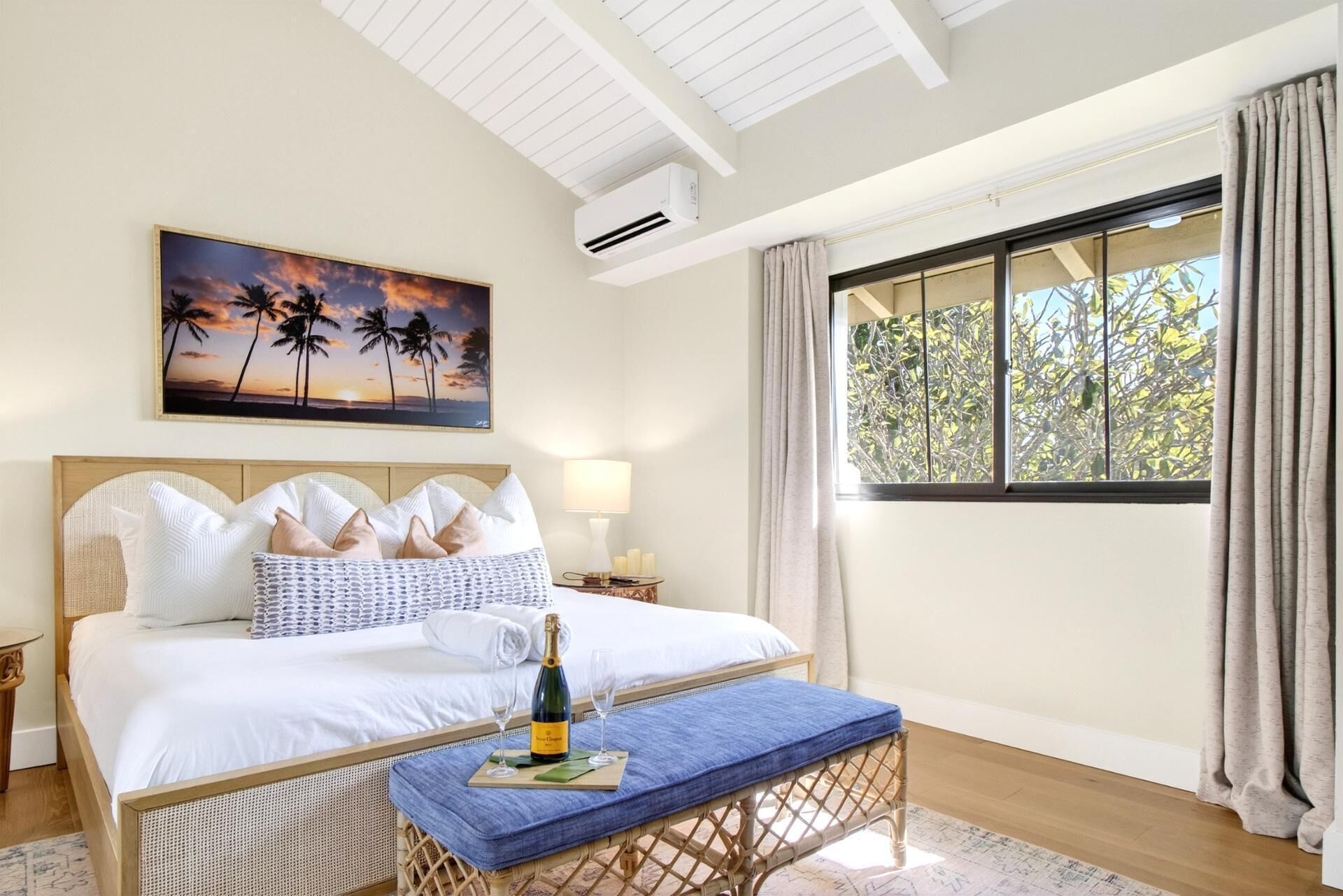 Una habitación de hotel con una cama, una mesita de noche, una ventana con cortinas, un aire acondicionado instalado en la pared y un cuadro de palmeras.