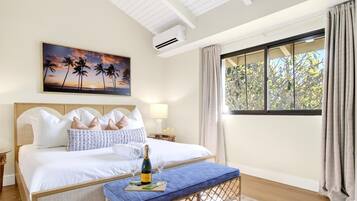 Una habitación de hotel con una cama, una mesita de noche, una ventana con cortinas, un aire acondicionado instalado en la pared y un cuadro de palmeras.