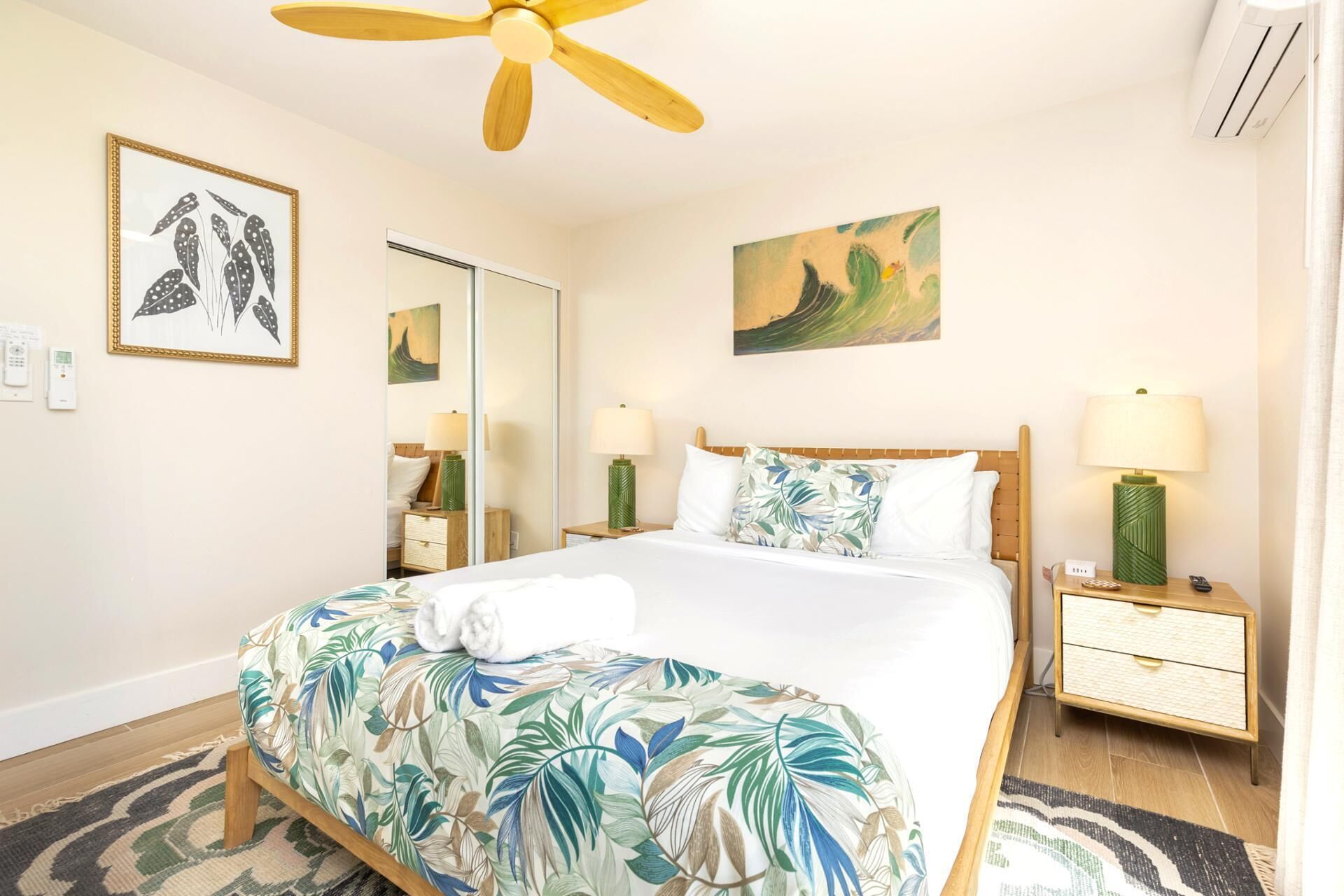 Ein Hotelzimmer mit einem Bett, zwei Nachttischen, einem Deckenventilator, einem Spiegel und Bildern an den Wänden.