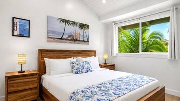 A bedroom with a wooden bed, white bedding, a beach-themed painting, and a window with a view of palm trees.