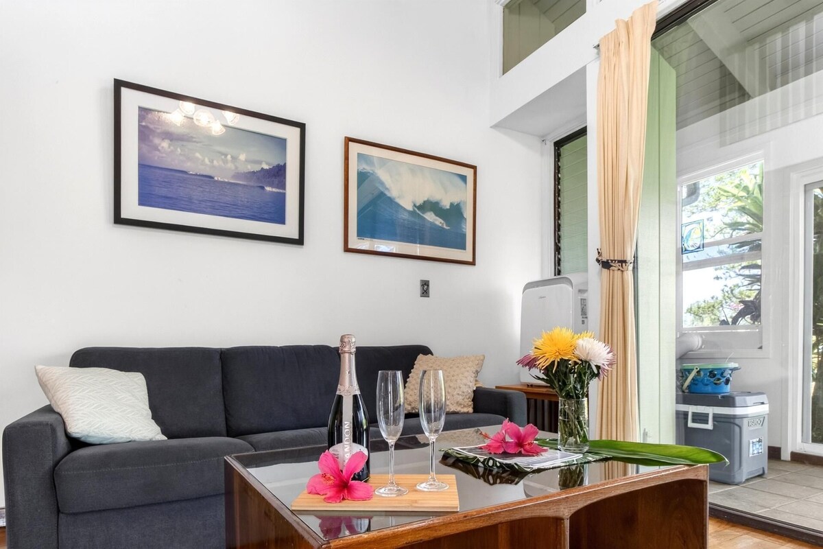 A living room with a dark grey sofa, two framed landscape paintings, a glass table with a bottle, two glasses, and a vase with flowers.
