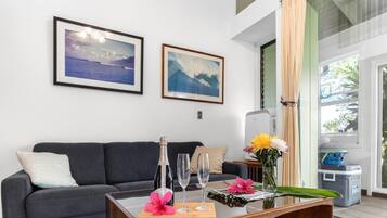 A living room with a dark grey sofa, two framed landscape paintings, a glass table with a bottle, two glasses, and a vase with flowers.
