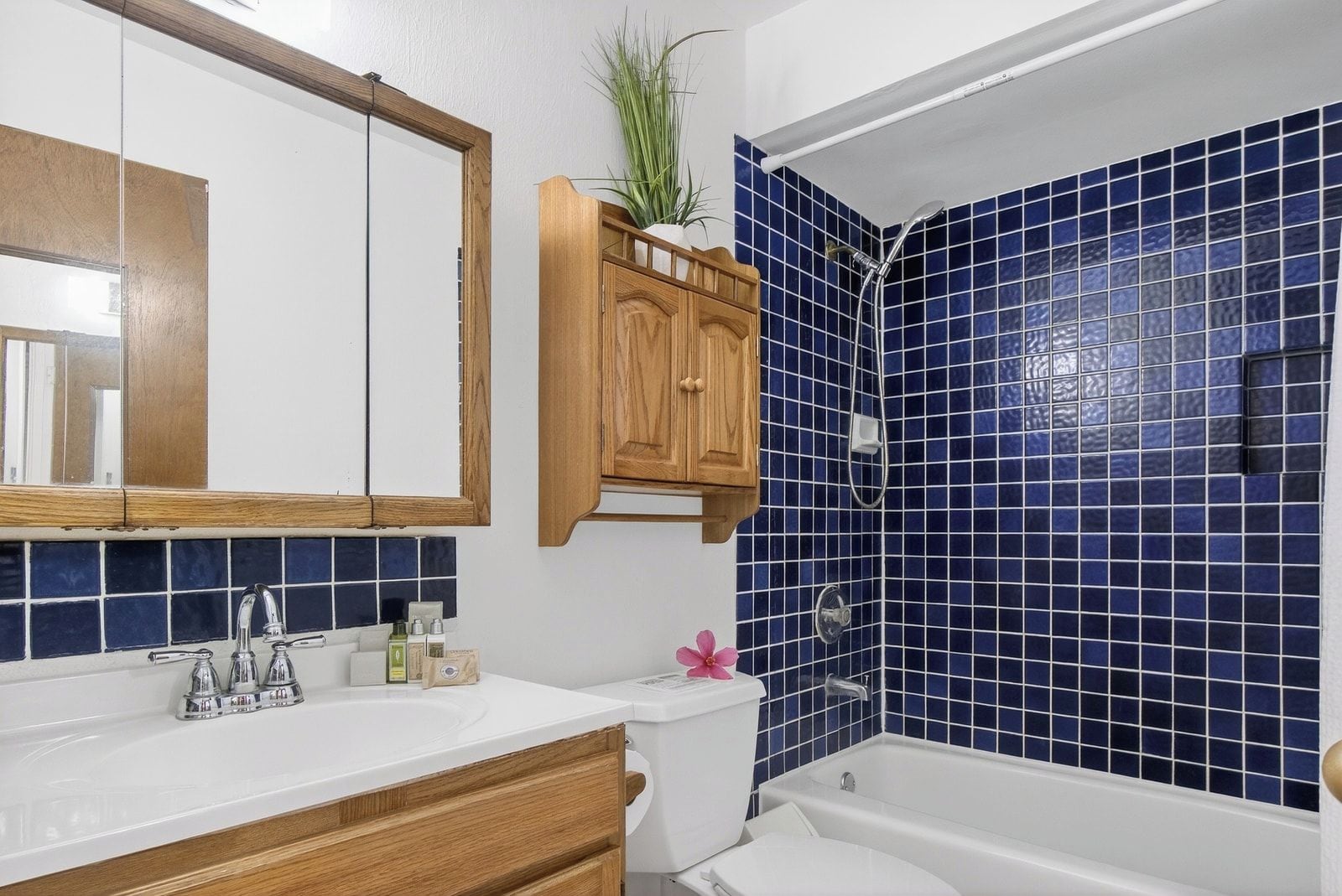 A bathroom with a white sink, wooden cabinets, a mirror, a shower with blue tiles, and a toilet.