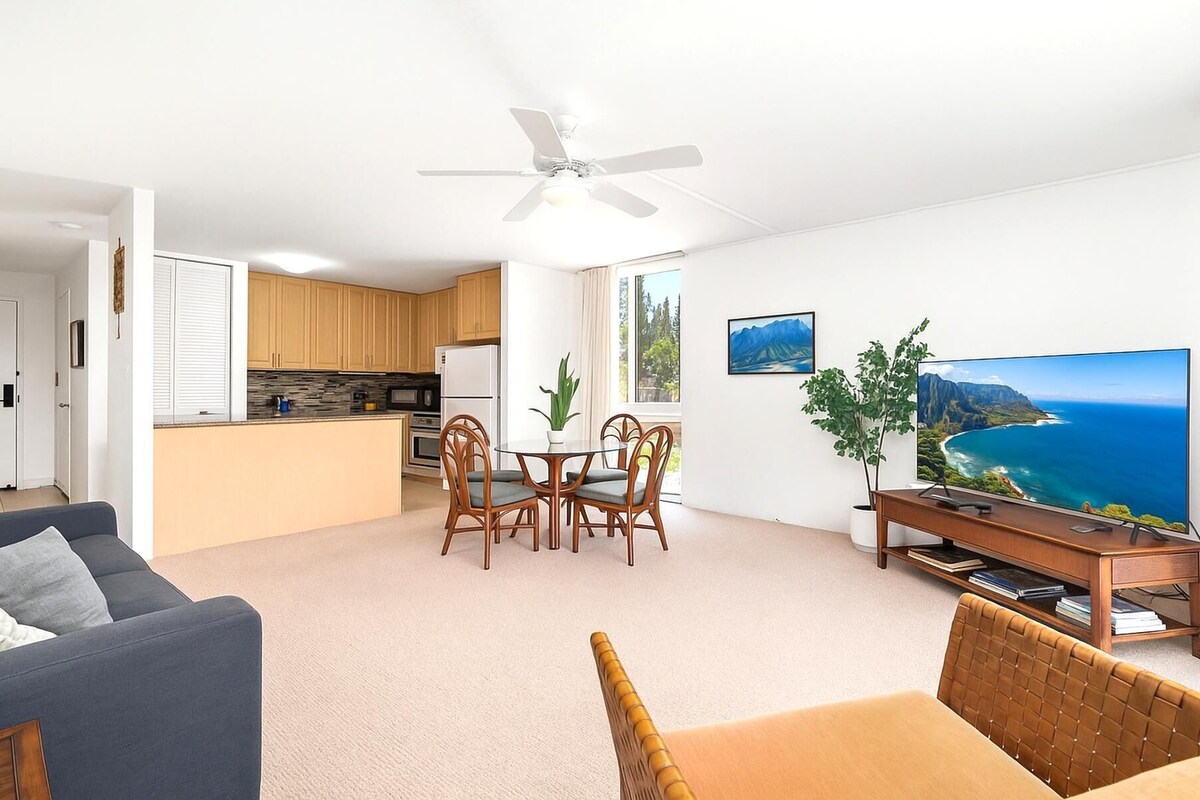 A modern living room with a sofa, a dining area with chairs, a kitchen with wooden cabinets, a ceiling fan, and a large TV displaying a scenic view.