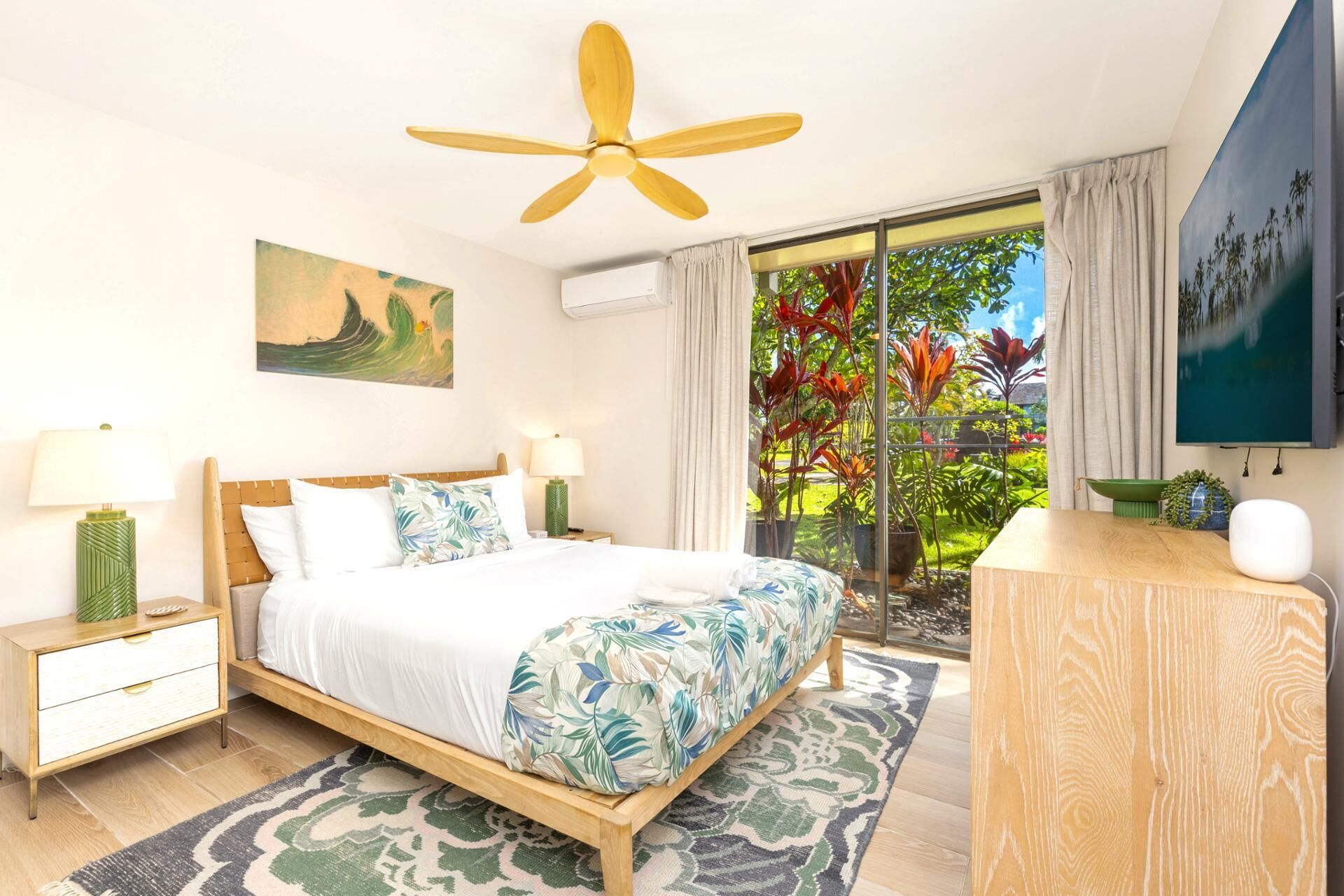 Ein Schlafzimmer mit Bett, Nachttischen, Deckenventilator, einem Gemälde, Blick auf einen Garten und einem Schreibtisch mit Stuhl.