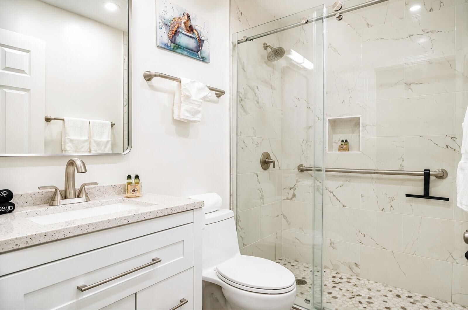 Un baño con ducha de vidrio, inodoro blanco, zona de ducha con azulejos de mármol y encimera con artículos de aseo.
