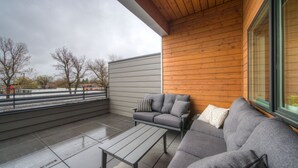 Ein überdachter Außenbereich mit einer grauen Couch und einem Couchtisch, Holzverkleidungen und Blick auf Bäume sowie ein Gebäude im Hintergrund.