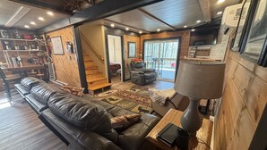 A cozy living room with a leather sofa, a wooden coffee table, and a lamp. There is a staircase leading to an upper level, and a large window providing a view of the outdoors. - Cabin in Hocking Hills with Hot Tub, Fire Pit and Covered Back Deck. (South Bloomingville)