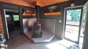 A cabin interior with a wood-burning stove, a kitchen area, and a door leading to an outdoor deck. - Cabin in Hocking Hills with Hot Tub, Fire Pit and Covered Back Deck. (South Bloomingville)
