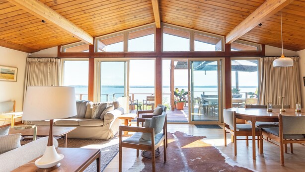 Un vaste espace de vie doté d’un haut plafond en bois, de grandes fenêtres offrant une vue sur la mer, un coin salon confortable avec un canapé et des fauteuils, ainsi qu’une table à manger avec des chaises.