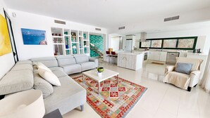 A modern living room with a grey sofa, a coffee table, and a colorful rug. There is a kitchen area with white cabinets and a window with blinds. - Villa Lili (El Perelló)