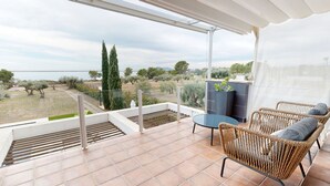 A balcony with a view of olive trees and a body of water, wicker furniture, and a small table. - Villa Lili (El Perelló)