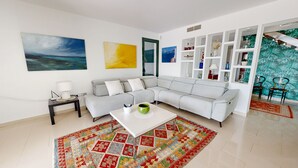 A modern living room with a grey sectional sofa, a white coffee table, and a colorful rug. There are abstract paintings on the walls and a patterned wall unit.