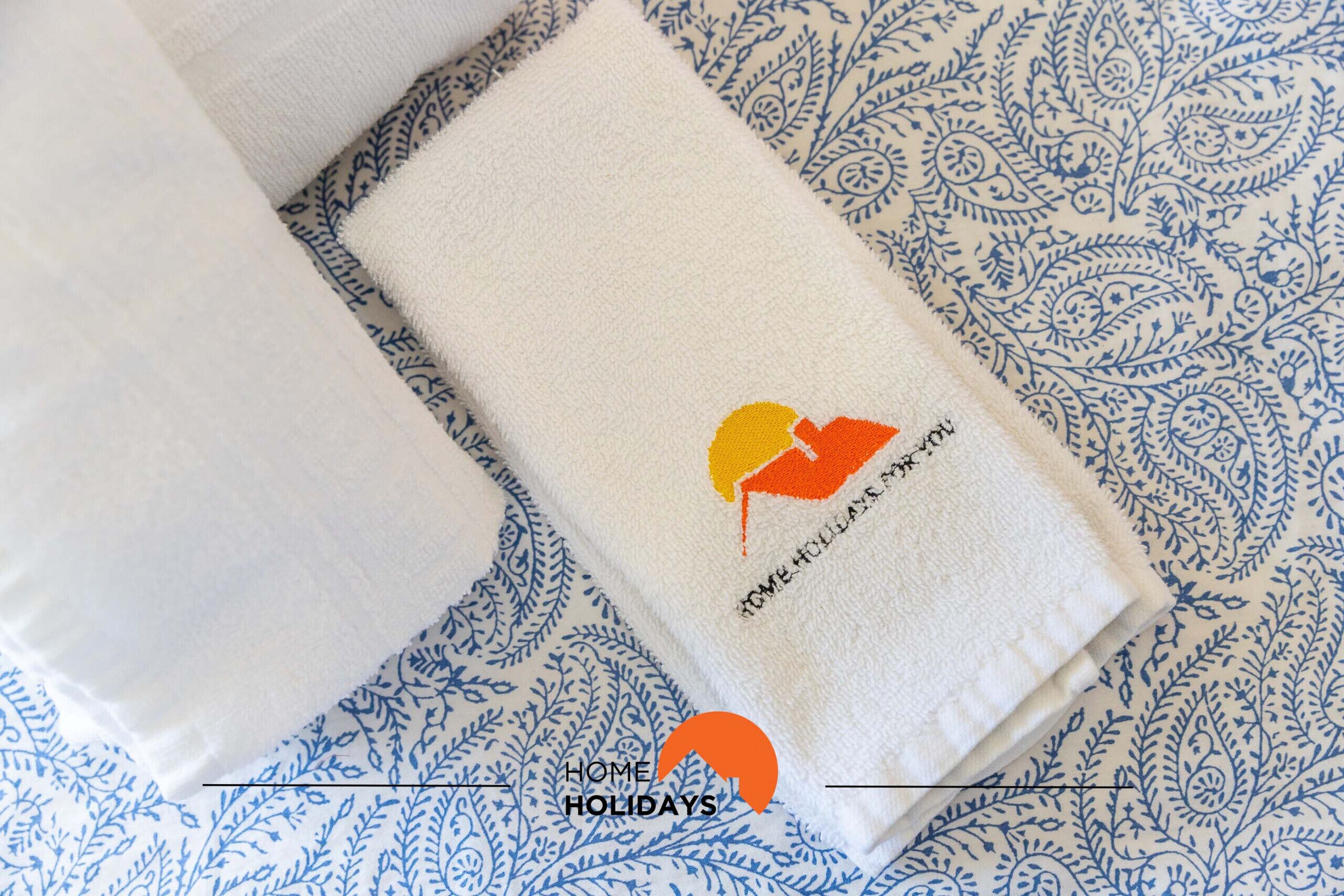 A neatly folded white towel with a logo and text on it, placed on a patterned blue and white bedspread.